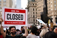 A large crowd of people gathers in a city setting, holding various signs. One prominent sign in the forefront reads 'Sorry, We're Closed Due to Climate Crisis #ClimateStrike.' The background features tall buildings and a black sculpture, suggesting an urban environment.