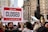 A large crowd of people gathers in a city setting, holding various signs. One prominent sign in the forefront reads 'Sorry, We're Closed Due to Climate Crisis #ClimateStrike.' The background features tall buildings and a black sculpture, suggesting an urban environment.