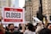 A large crowd of people gathers in a city setting, holding various signs. One prominent sign in the forefront reads 'Sorry, We're Closed Due to Climate Crisis #ClimateStrike.' The background features tall buildings and a black sculpture, suggesting an urban environment.