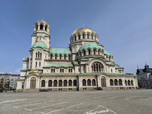 A large, ornate cathedral with multiple domes, featuring a combination of green and gold accents atop a white stone structure. The architecture exhibits a blend of Romanesque and Neo-Byzantine styles. It is situated in a spacious, paved square with additional buildings visible in the background.