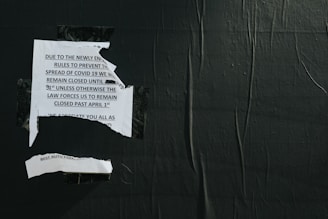 A torn notice is taped to a dark, textured wall. The message on the notice pertains to COVID-19 closure regulations and dates. The paper appears ripped, suggesting wear or destruction.