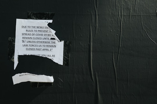A torn notice is taped to a dark, textured wall. The message on the notice pertains to COVID-19 closure regulations and dates. The paper appears ripped, suggesting wear or destruction.