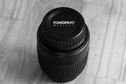 A camera lens with a cap featuring the brand name on a wooden surface. The lens is cylindrical and appears in monochrome.