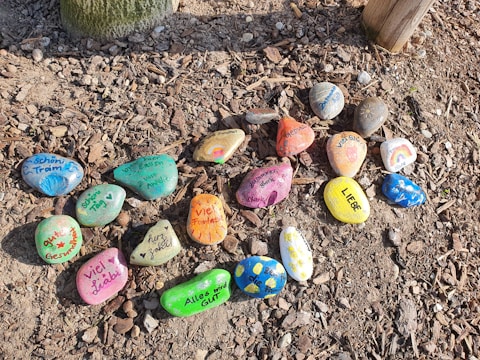 A collection of colorful, painted stones arranged on a bed of gravel or soil. Each stone has a unique design, featuring positive messages, patterns, or symbols in various colors. Some stones have words in a foreign language, while others depict simple, cheerful drawings. The stones are scattered in a somewhat organized pattern.