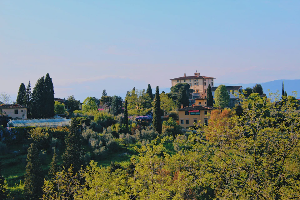 The image depicts a picturesque landscape featuring a large, multi-story building with a red roof and white walls nestled amidst lush greenery. The building is surrounded by an array of trees and shrubs, creating a harmonious blend of architecture and nature. In the distance, mountains can be seen under a clear blue sky, adding depth to the scene.