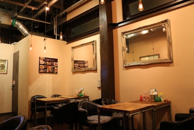 A cozy restaurant interior features wooden tables and black chairs set against cream-colored walls. Mirrors are mounted on the wall, reflecting the warm lighting from exposed bulbs hanging from the ceiling. A menu board displays detailed images of food options.