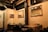 A cozy restaurant interior features wooden tables and black chairs set against cream-colored walls. Mirrors are mounted on the wall, reflecting the warm lighting from exposed bulbs hanging from the ceiling. A menu board displays detailed images of food options.