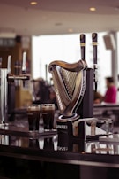 A bar counter features a prominent Guinness beer tap shaped like a harp with two draft levers, branded with the Guinness logo. Two pints of dark stout beer sit on the shiny black counter, showcasing the brand's signature look. The blurred background includes soft lighting and a figure in casual attire.