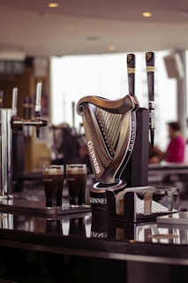 A bar counter features a prominent Guinness beer tap shaped like a harp with two draft levers, branded with the Guinness logo. Two pints of dark stout beer sit on the shiny black counter, showcasing the brand's signature look. The blurred background includes soft lighting and a figure in casual attire.