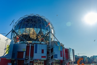 A geodesic dome structure with a reflective surface is situated above a building featuring various colors and geometric designs. The sky is clear with a few birds flying, and the sun is shining brightly, creating a lens flare. The setting appears urban, with additional buildings and construction cranes visible in the background.