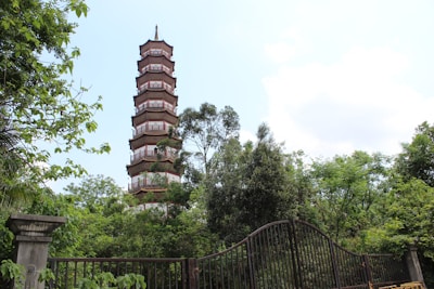 A tiered pagoda tower rises above lush green foliage with partly cloudy skies in the background. A wrought iron gate is in the foreground, partly obscured by the plants and trees.