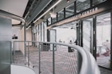A modern interior space of an entrepreneurship center with large glass windows and open doors. The room contains people sitting at tables, engaging in various activities. The sleek design features metal railings and exposed ductwork in the ceiling, creating an industrial feel.