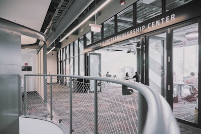 A modern interior space of an entrepreneurship center with large glass windows and open doors. The room contains people sitting at tables, engaging in various activities. The sleek design features metal railings and exposed ductwork in the ceiling, creating an industrial feel.