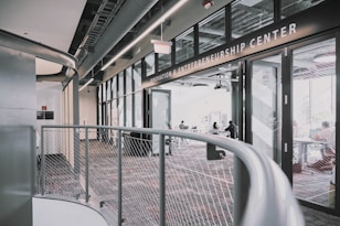A modern interior space of an entrepreneurship center with large glass windows and open doors. The room contains people sitting at tables, engaging in various activities. The sleek design features metal railings and exposed ductwork in the ceiling, creating an industrial feel.