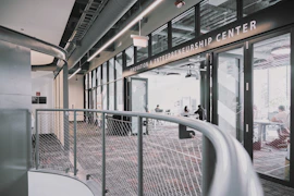 A modern interior space of an entrepreneurship center with large glass windows and open doors. The room contains people sitting at tables, engaging in various activities. The sleek design features metal railings and exposed ductwork in the ceiling, creating an industrial feel.