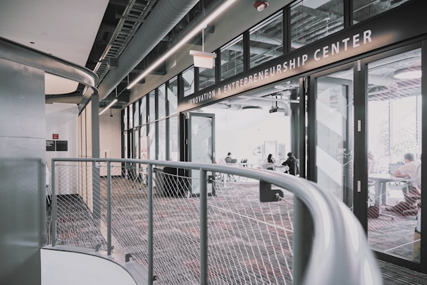 A modern interior space of an entrepreneurship center with large glass windows and open doors. The room contains people sitting at tables, engaging in various activities. The sleek design features metal railings and exposed ductwork in the ceiling, creating an industrial feel.
