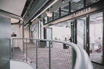 A modern interior space of an entrepreneurship center with large glass windows and open doors. The room contains people sitting at tables, engaging in various activities. The sleek design features metal railings and exposed ductwork in the ceiling, creating an industrial feel.