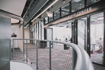 A modern interior space of an entrepreneurship center with large glass windows and open doors. The room contains people sitting at tables, engaging in various activities. The sleek design features metal railings and exposed ductwork in the ceiling, creating an industrial feel.
