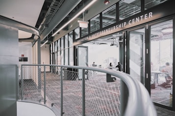 A modern interior space of an entrepreneurship center with large glass windows and open doors. The room contains people sitting at tables, engaging in various activities. The sleek design features metal railings and exposed ductwork in the ceiling, creating an industrial feel.