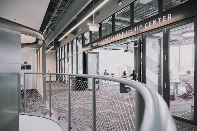 A modern interior space of an entrepreneurship center with large glass windows and open doors. The room contains people sitting at tables, engaging in various activities. The sleek design features metal railings and exposed ductwork in the ceiling, creating an industrial feel.