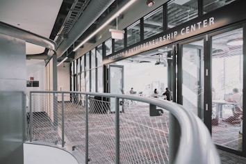 A modern interior space of an entrepreneurship center with large glass windows and open doors. The room contains people sitting at tables, engaging in various activities. The sleek design features metal railings and exposed ductwork in the ceiling, creating an industrial feel.