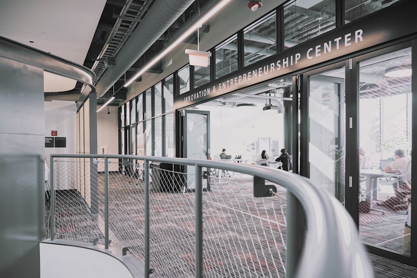 A modern interior space of an entrepreneurship center with large glass windows and open doors. The room contains people sitting at tables, engaging in various activities. The sleek design features metal railings and exposed ductwork in the ceiling, creating an industrial feel.