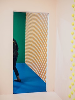 A colorful interior scene featuring a view through a doorway. The floor is a vivid blue, and the walls are painted with diagonal yellow and white stripes. A person wearing jeans and a dark jacket is partially visible, with only part of their body seen as they walk through the doorway. Yellow objects are attached to a white wall on the right side of the image.