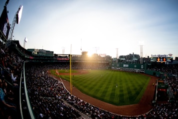 A large baseball stadium filled with spectators, featuring a bright, well-maintained field. The sun sets in the distance, casting a warm glow over the scene. The stadium's seating areas are packed with people, creating a lively atmosphere, while the structures around the field display advertisements and logos.