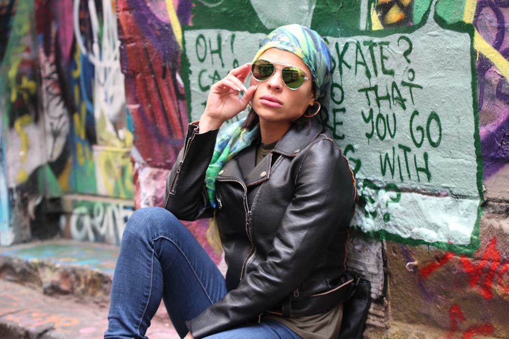 woman in black leather jacket and blue denim jeans wearing green knit cap sitting on concrete