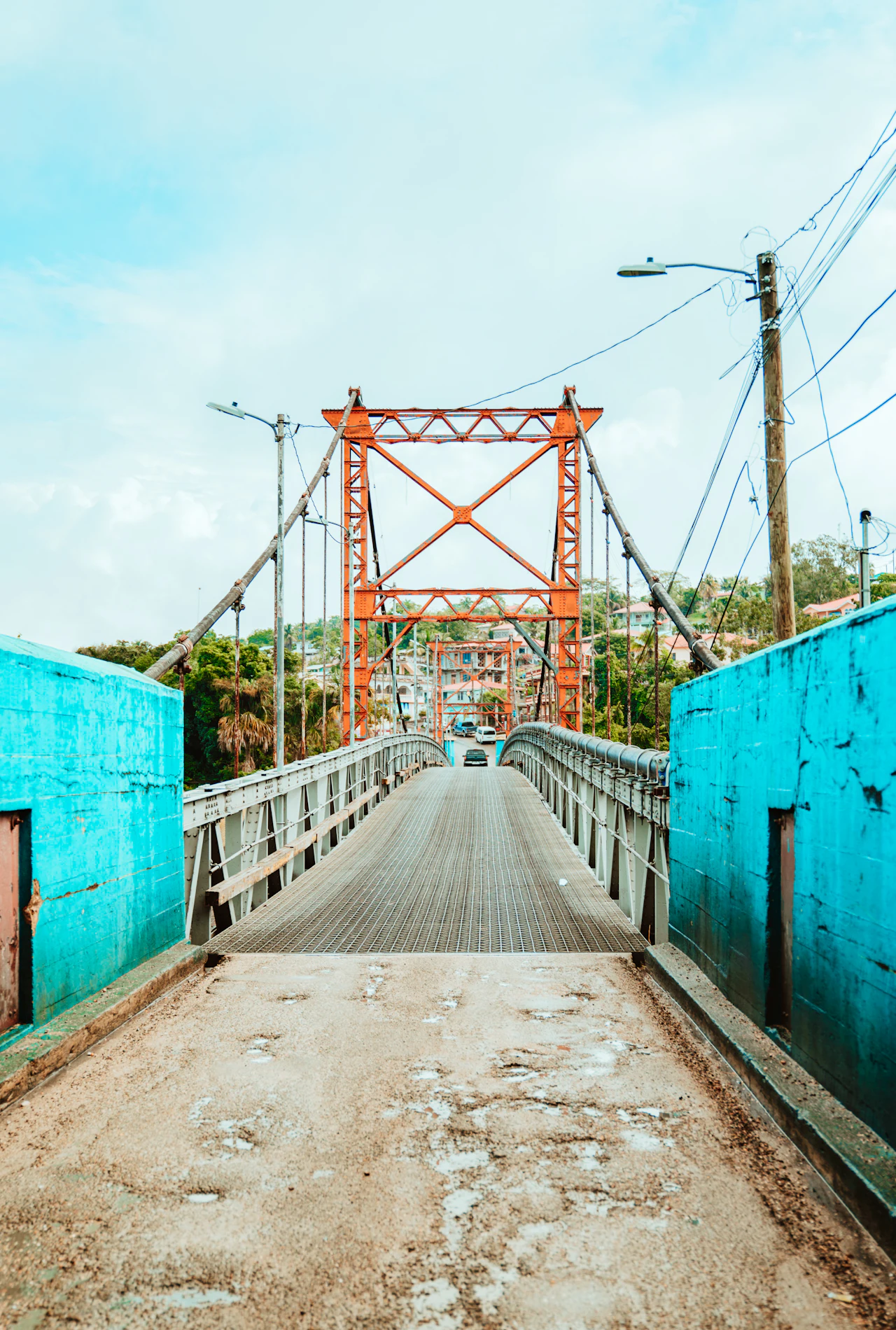 Looking along a narrow road onto an orange-painted suspension bridge, with turquoise walls on either side.