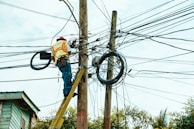 A worker is on a ladder fixing a complex network of electrical wires on a utility pole. The pole is surrounded by numerous tangled cables. The worker is wearing a hard hat and reflective vest, indicating safety precautions. Parts of a building and trees are visible in the background.