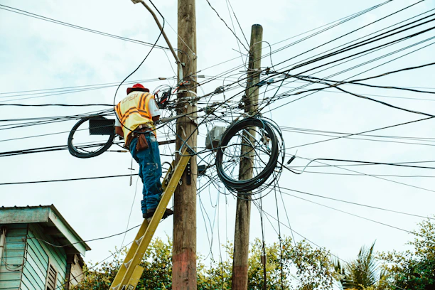 A worker is on a ladder fixing a complex network of electrical wires on a utility pole. The pole is surrounded by numerous tangled cables. The worker is wearing a hard hat and reflective vest, indicating safety precautions. Parts of a building and trees are visible in the background.