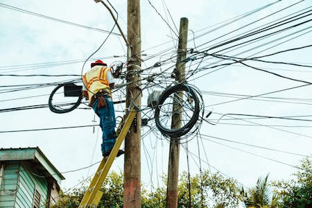 A worker is on a ladder fixing a complex network of electrical wires on a utility pole. The pole is surrounded by numerous tangled cables. The worker is wearing a hard hat and reflective vest, indicating safety precautions. Parts of a building and trees are visible in the background.