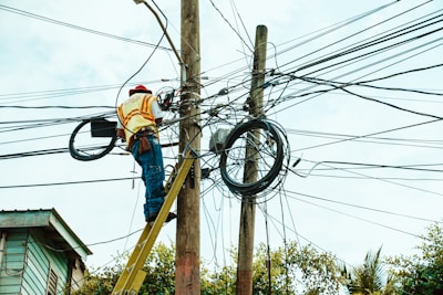 A worker is on a ladder fixing a complex network of electrical wires on a utility pole. The pole is surrounded by numerous tangled cables. The worker is wearing a hard hat and reflective vest, indicating safety precautions. Parts of a building and trees are visible in the background.