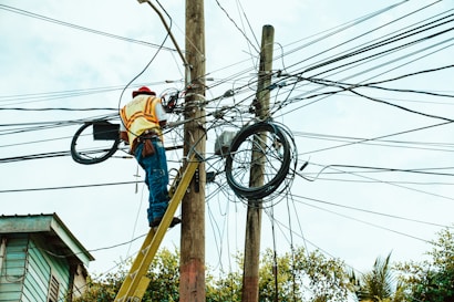 A worker is on a ladder fixing a complex network of electrical wires on a utility pole. The pole is surrounded by numerous tangled cables. The worker is wearing a hard hat and reflective vest, indicating safety precautions. Parts of a building and trees are visible in the background.