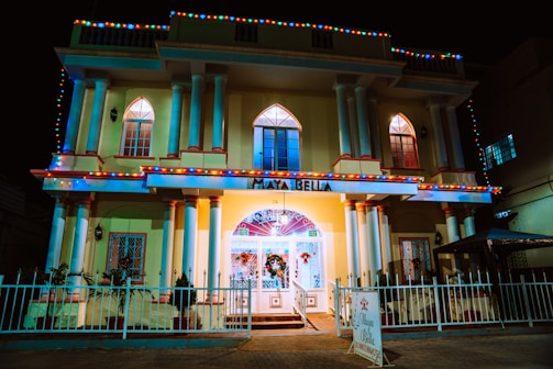 A two-story building with a grand entrance, featuring tall columns and multiple arched windows. The facade is decorated with colorful string lights, casting a festive glow. The sign above the entrance reads 'Maya Bella'. There is a white fence with decorative wrought iron surrounding the property.