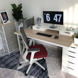 A modern, well-organized workspace with a wooden desk featuring a dual monitor setup displaying the time. A black keyboard and mouse are neatly placed on the desk, with a toy figure and a decorative plant nearby. An ergonomic chair with a red seat is positioned in front of the desk. A standing shelf in the corner holds a framed picture, and there's a large leafy plant.