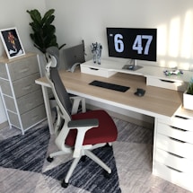 A modern, well-organized workspace with a wooden desk featuring a dual monitor setup displaying the time. A black keyboard and mouse are neatly placed on the desk, with a toy figure and a decorative plant nearby. An ergonomic chair with a red seat is positioned in front of the desk. A standing shelf in the corner holds a framed picture, and there's a large leafy plant.
