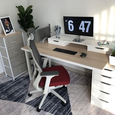 A modern, well-organized workspace with a wooden desk featuring a dual monitor setup displaying the time. A black keyboard and mouse are neatly placed on the desk, with a toy figure and a decorative plant nearby. An ergonomic chair with a red seat is positioned in front of the desk. A standing shelf in the corner holds a framed picture, and there's a large leafy plant.