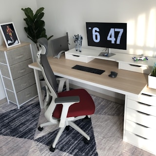 A modern, well-organized workspace with a wooden desk featuring a dual monitor setup displaying the time. A black keyboard and mouse are neatly placed on the desk, with a toy figure and a decorative plant nearby. An ergonomic chair with a red seat is positioned in front of the desk. A standing shelf in the corner holds a framed picture, and there's a large leafy plant.