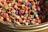 A variety of colorful dried flowers and petals are clustered together inside a container. The flowers exhibit diverse hues, including orange, pink, purple, green, and cream. The texture is crinkled and layered, and the composition creates a vibrant mosaic of colors with an organic feel.