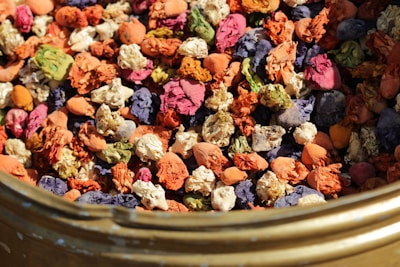 A variety of colorful dried flowers and petals are clustered together inside a container. The flowers exhibit diverse hues, including orange, pink, purple, green, and cream. The texture is crinkled and layered, and the composition creates a vibrant mosaic of colors with an organic feel.