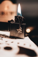 A lit lighter with a blue and orange flame rests on a table. The lighter has a dangling metallic angel charm. Below it, out of focus, is a sheet of paper with markings that resemble musical notes.