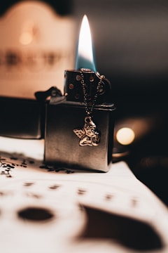 A lit lighter with a blue and orange flame rests on a table. The lighter has a dangling metallic angel charm. Below it, out of focus, is a sheet of paper with markings that resemble musical notes.