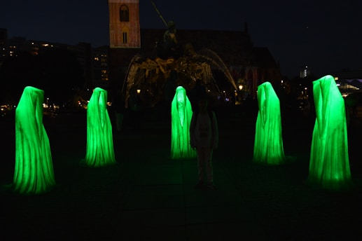 Six tall, ghost-like figures in glowing green robes stand in a circle in a dark outdoor setting. Their forms are illuminated, creating an eerie atmosphere. In the background, a large sculpture and a building are visible.