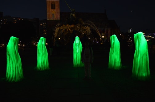 Six tall, ghost-like figures in glowing green robes stand in a circle in a dark outdoor setting. Their forms are illuminated, creating an eerie atmosphere. In the background, a large sculpture and a building are visible.