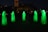 Six tall, ghost-like figures in glowing green robes stand in a circle in a dark outdoor setting. Their forms are illuminated, creating an eerie atmosphere. In the background, a large sculpture and a building are visible.