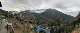 A mountainous landscape with lush green hills. The hills are covered with a mix of dense forests and small villages, featuring multi-story houses. A mist covers parts of the hills, creating a serene atmosphere. The sky is overcast, adding to the tranquil mood. Nearby, a paved path and several parked cars can be seen.