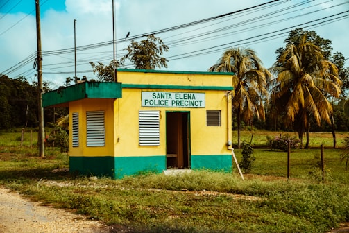 A small yellow and green building labeled 'Santa Elena Police Precinct' is surrounded by greenery and palm trees. The structure has two windows with shutters and a doorway that is partially open. Power lines run across the scene, and a dirt path leads up to the building.