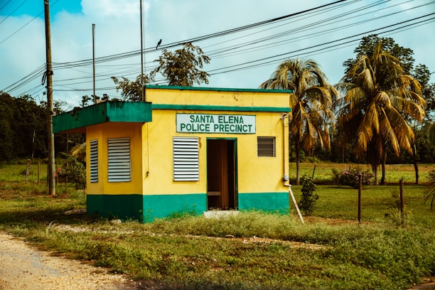 A small yellow and green building labeled 'Santa Elena Police Precinct' is surrounded by greenery and palm trees. The structure has two windows with shutters and a doorway that is partially open. Power lines run across the scene, and a dirt path leads up to the building.